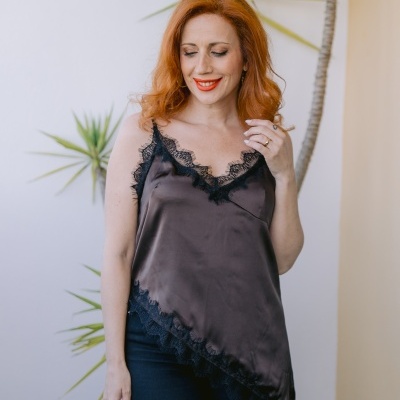 Mulher com blusa acetinada castanha escuro com renda preta e calças pretas, planta ao fundo