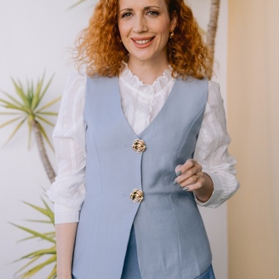 mulher de cabelo ruivo usa colete azul claro com botões dourados florais e blusa branca com gola babada