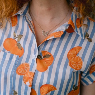 Camisa azul e branca com laranjas, colar de borboleta dourado e brincos de argola dourados