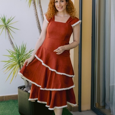 Mulher com vestido terracota de bainhas brancas, em ambiente exterior perto de plantas.