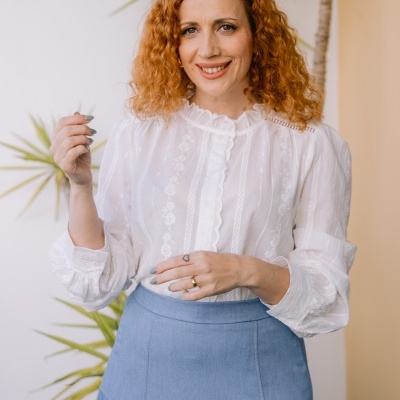 Mulher com blusa branca de manga comprida e saia azul clara em ambiente interior com planta