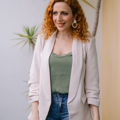 Mulher com blusa verde, blazer bege e jeans azul junto a plantas