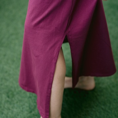Saia comprida roxo vinho com fenda lateral em tecido algodão, vista de perto com pés descalços sobre relva sintética verde.