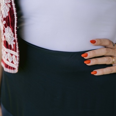 Detalhe de vestido preto com blusa branca e bordado vermelho, mão com unhas vermelhas e anel