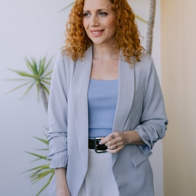 Mulher com blazer cinza claro e calças brancas num ambiente com plantas.