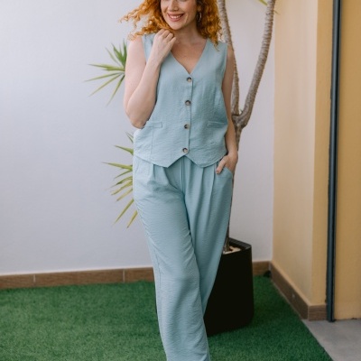Conjunto feminino azul claro com colete e calças largas, sapatos dourados, planta e parede de fundo