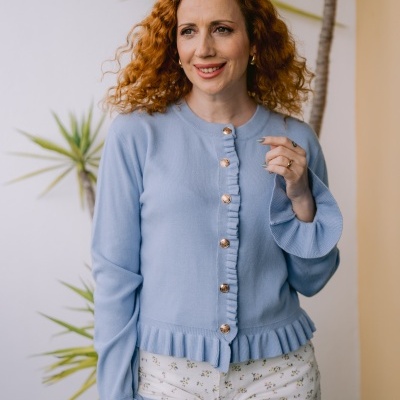 Mulher a usar casaco azul claro com botões dourados e calças brancas florais, com planta ao fundo