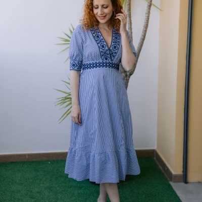 Mulher com vestido azul claro às riscas brancas e bordados azuis, sapatos dourados, em relva artificial junto a parede.