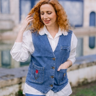 Mulher ruiva com colete de ganga azul e camisa branca em cenário exterior com lago e azulejos.