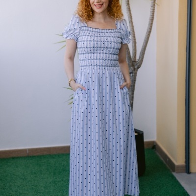 Mulher com vestido comprido azul claro às riscas e bolinhas azuis em ambiente exterior com relva e planta.