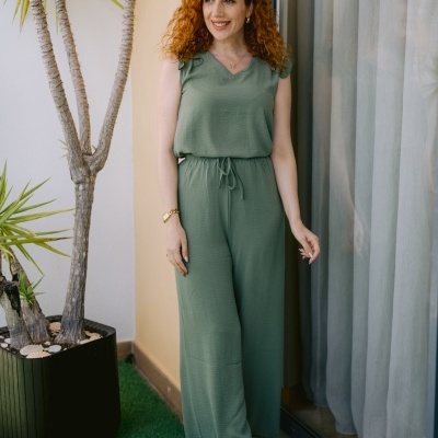 Mulher com macacão verde claro ao lado de planta em vaso e janela com cortina