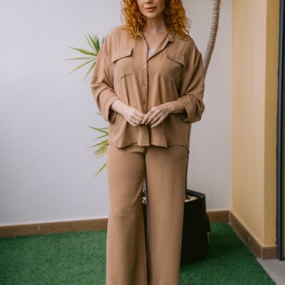 Mulher com camisa bege e calças largas bege, botas douradas, em ambiente com parede branca e planta.