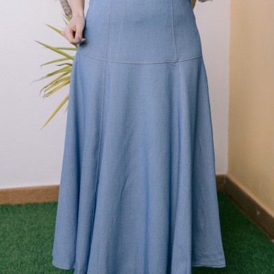 Saia comprida azul clara em denim com camisa branca e plantas ao fundo