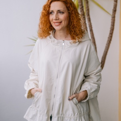 Mulher com casaco bege claro de capuz e fecho de correr ao ar livre junto a planta