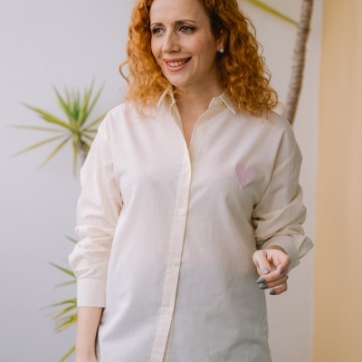 Mulher com camisa às riscas amarelas claras com coração rosa e calças jeans azuis num ambiente com plantas.