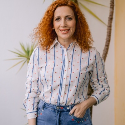 Mulher com camisa branca com padrão de corações vermelhos e linhas azuis, usando jeans azul claro com bordados, em ambiente com planta ao fundo