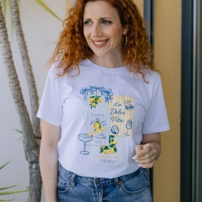 Mulher com t-shirt branca estampada com limões e textos em azul e amarelo, jeans azuis e cabelos ruivos encaracolados.