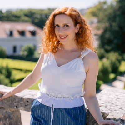 Mulher com top branco e calças às riscas azuis encostada num muro de pedra ao ar livre