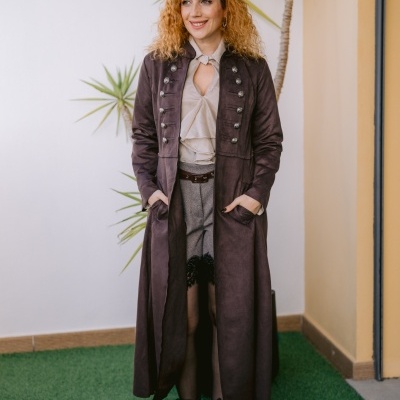 Mulher com casaco castanho comprido, blusa bege, calções cinza com renda preta e botas pretas