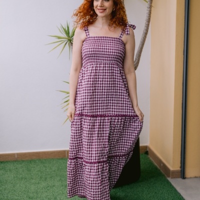 Mulher com vestido às quadrículas bordeaux e branco num ambiente exterior com planta e relva artificial.