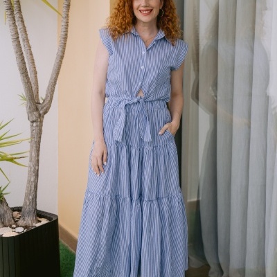 Vestido comprido azul claro às riscas brancas com cinto, usado por mulher junto a porta e planta