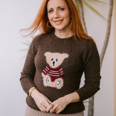 Mulher ruiva com camisola de malha castanha com urso e saia bege às riscas numa varanda com planta