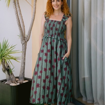Vestido longo verde com flores vermelhas usado por mulher junto a planta e janela