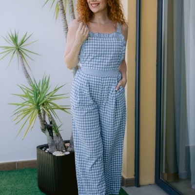 Mulher com macacão azul e branco xadrez junto a planta em vaso e parede amarela