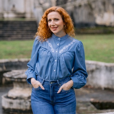 Mulher com camisa azul bordada e jeans junto a uma fonte de pedra num jardim.