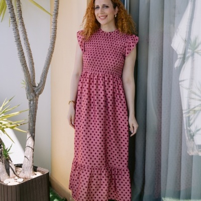 Mulher com vestido rosa de bolinhas roxas perto de planta e janela com cortinas
