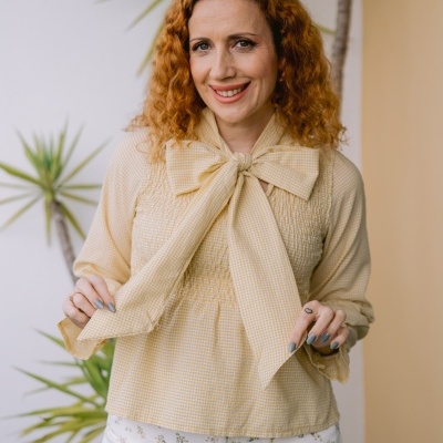 Mulher com blusa amarela de laço e calças brancas florais junto a plantas