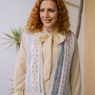 Mulher com blusa amarela de manga comprida, colete patchwork e calças florais brancas.