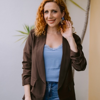 Mulher com blazer castanho, blusa azul clara e calças de ganga junto a plantas e parede branca e amarela.