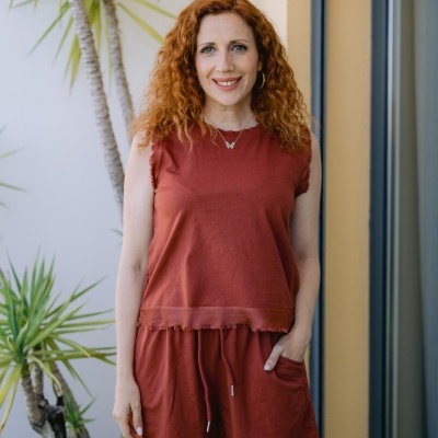 Mulher de cabelo ruivo com roupa castanha a sorrir numa varanda com plantas.