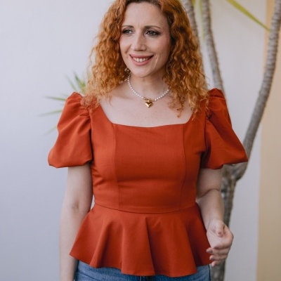 Mulher ruiva com blusa laranja de mangas bufantes e jeans azul claro com planta ao fundo