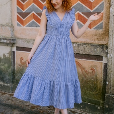 Vestido midi azul às riscas com folhos e botões, sapatos dourados e parede decorada ao fundo