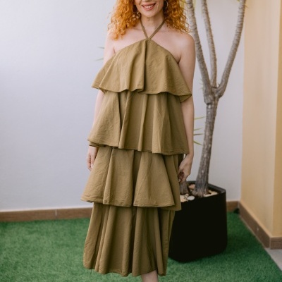 Mulher com vestido bege e sapatos dourados junto a planta em vaso.