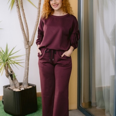 Mulher de camisa e calças bordô com franjas nas mangas, sapatos dourados com atacadores azuis, em varanda com planta.