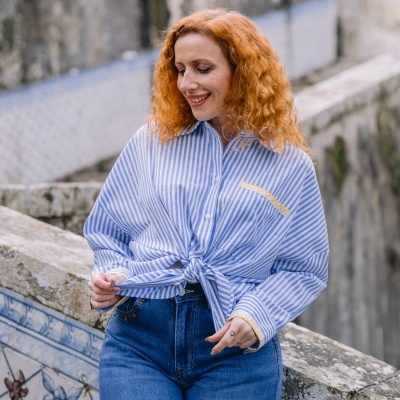 Mulher com camisa azul e branca às riscas e calças de ganga azuis junto a parede com azulejos