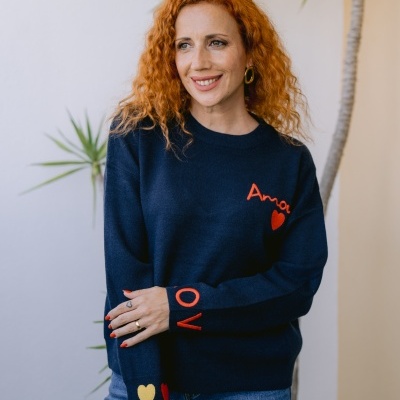 Mulher com camisola azul escura com palavras Amor e LOVE em vermelho e amarelo, jeans azuis, planta ao fundo