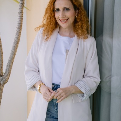 Mulher com casaco branco e t-shirt branca junto a janela com cortinado cinzento