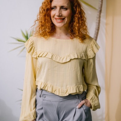 Mulher a usar blusa amarela com folhos e calças cinza em ambiente interior com plantas