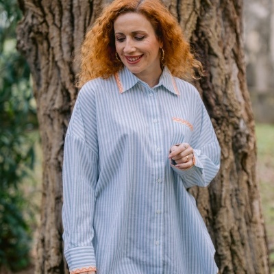 Camisa feminina às riscas azul claro e branco com bordados laranja e calças azuis junto a uma árvore