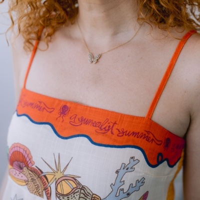 Vestido branco com estampado marinho e alças laranja, detalhes em texto e ilustrações de conchas e corais