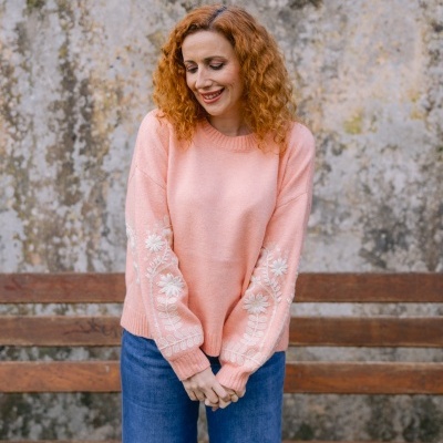 Mulher de vermelho com camisola rosa bordada e calças de ganga junto a banco de madeira