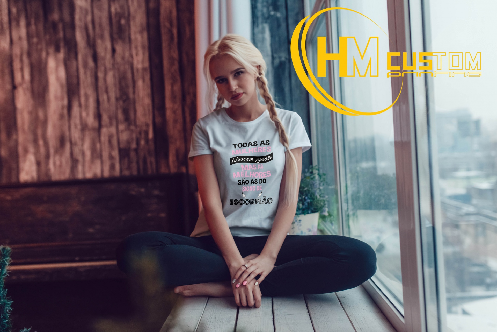 T-Shirt "As melhores Mulheres são do Signo de..." Mulher loira sentada junto a uma janela grande com t-shirt branca com texto em português