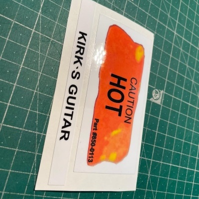 Etiqueta autocolante para guitarra com texto 'KIRK'S GUITAR' e etiqueta 'CAUTION HOT' sobre superfície verde de corte.