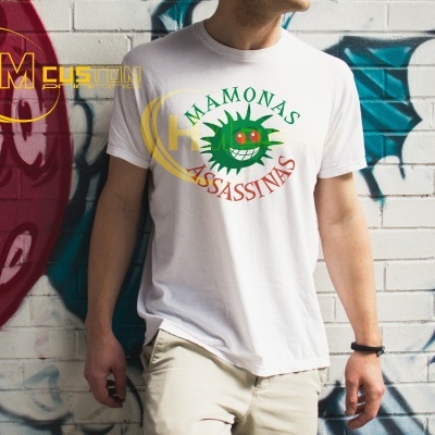 Pessoa veste uma T-shirt branca com estampa colorida 'MAMONAS ASSASSINAS' junto a uma parede grafitada.