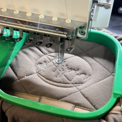 Máquina de bordar com aro verde bordando tecido cinza acolchoado com padrão de losangos e círculo com texto