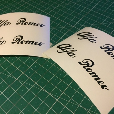 Folhas de papel com autocolantes 'Alfa Romeo' em preto sobre tapete de corte verde.